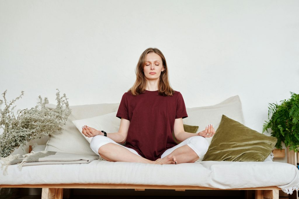 How Blood Sugar And Hormones Work Together To Influence Women’s Health Woman meditating indoors on a sofa, exuding calmness and mindfulness, surrounded by greenery.